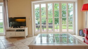 Quarto com televisão sobre um armário branco, mesa com tampo de vidro e uma janela grande com vista para móveis de exterior.