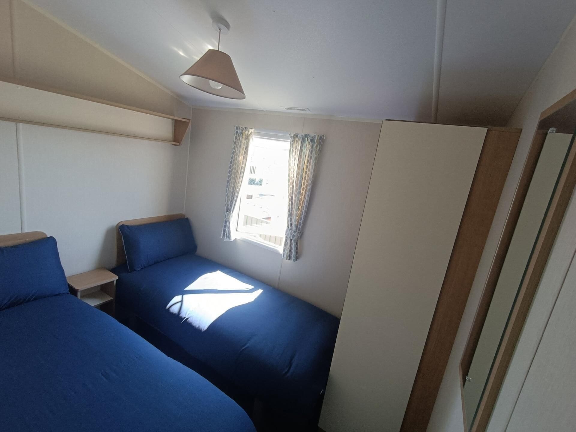Quarto pequeno com duas camas, janela com cortinas, uma luminária pendente e uma porta.