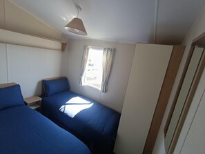 A compact room with two beds, a window with curtains, a hanging lamp, and a door. - Cozy 2 Bedroom Caravan Golden Sands (Rhyl)