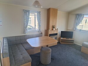 A compact living space with a sofa, a small table, a television, and a window with curtains. - Cozy 2 Bedroom Caravan Golden Sands (Rhyl)