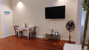 A living room with a flat-screen TV, a table with chairs, a fan, and a potted plant.