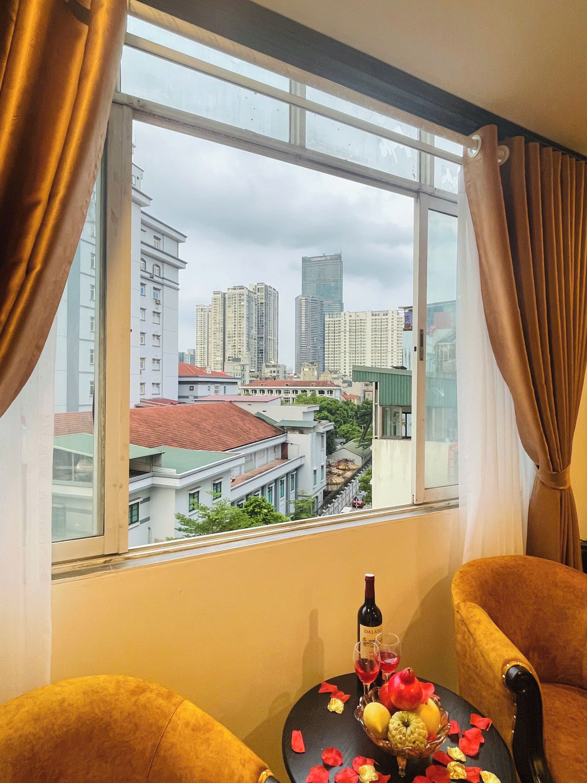 A room with a view of a cityscape through a large window, a table with a bottle and glasses, and two chairs.