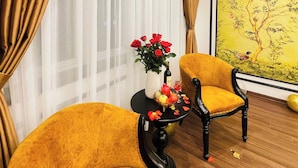 A room with a wooden floor, a yellow armchair, a table with a vase of red flowers, and a wall with a framed painting. - Vera Luxury Hotel (Hanoi)