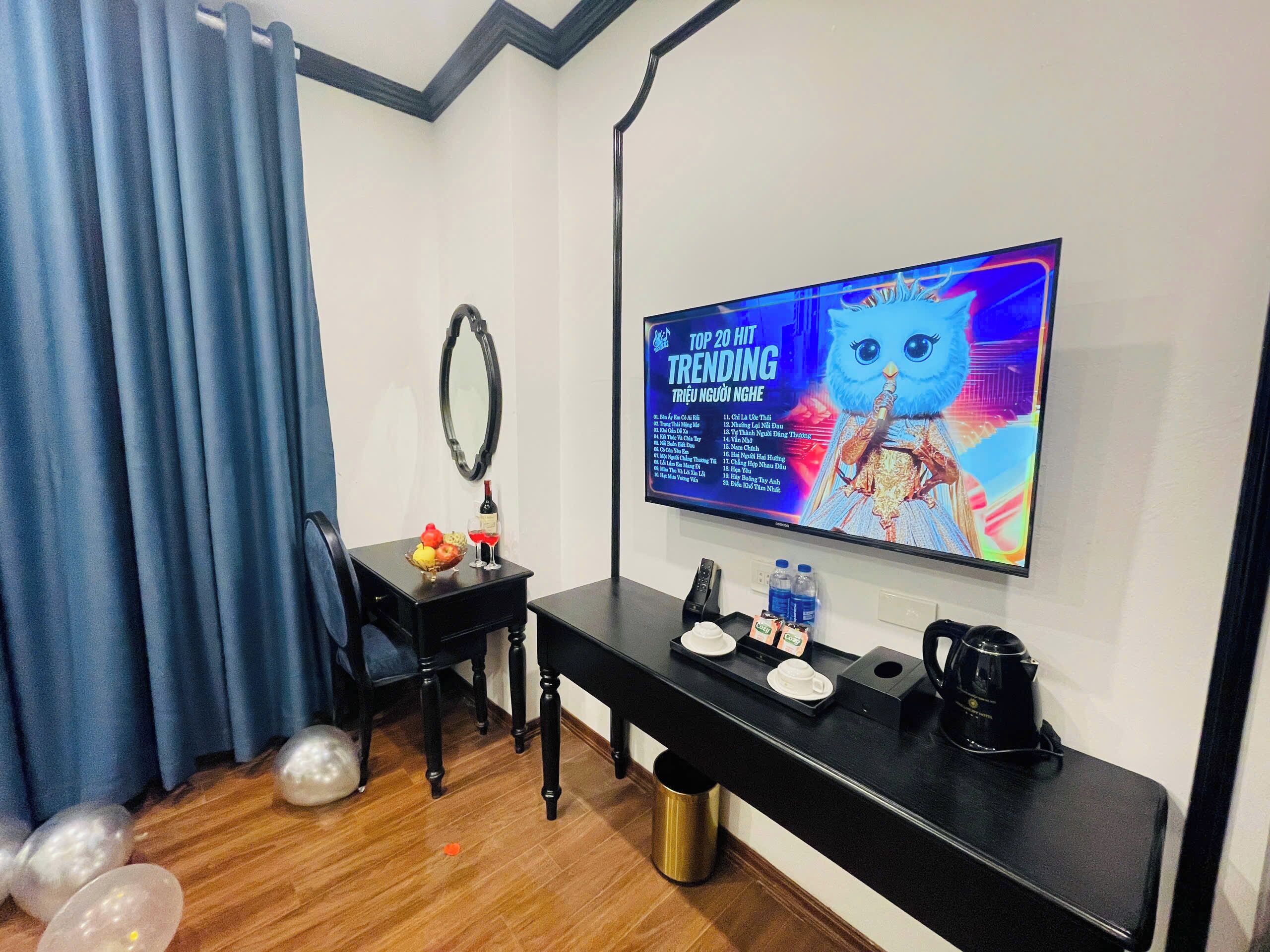 Habitación de hotel con televisor de pantalla plana mostrando una lista de éxitos musicales, una pequeña mesa con una botella y un vaso, una silla y una consola negra con cafetera.