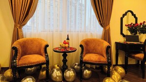 A room with two upholstered chairs, a small round table with a bottle and fruit, and a mirror on the wall. - Vera Luxury Hotel (Hanoi)