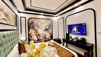 A hotel room with a large bed, a television, and a wall-mounted screen displaying a program.