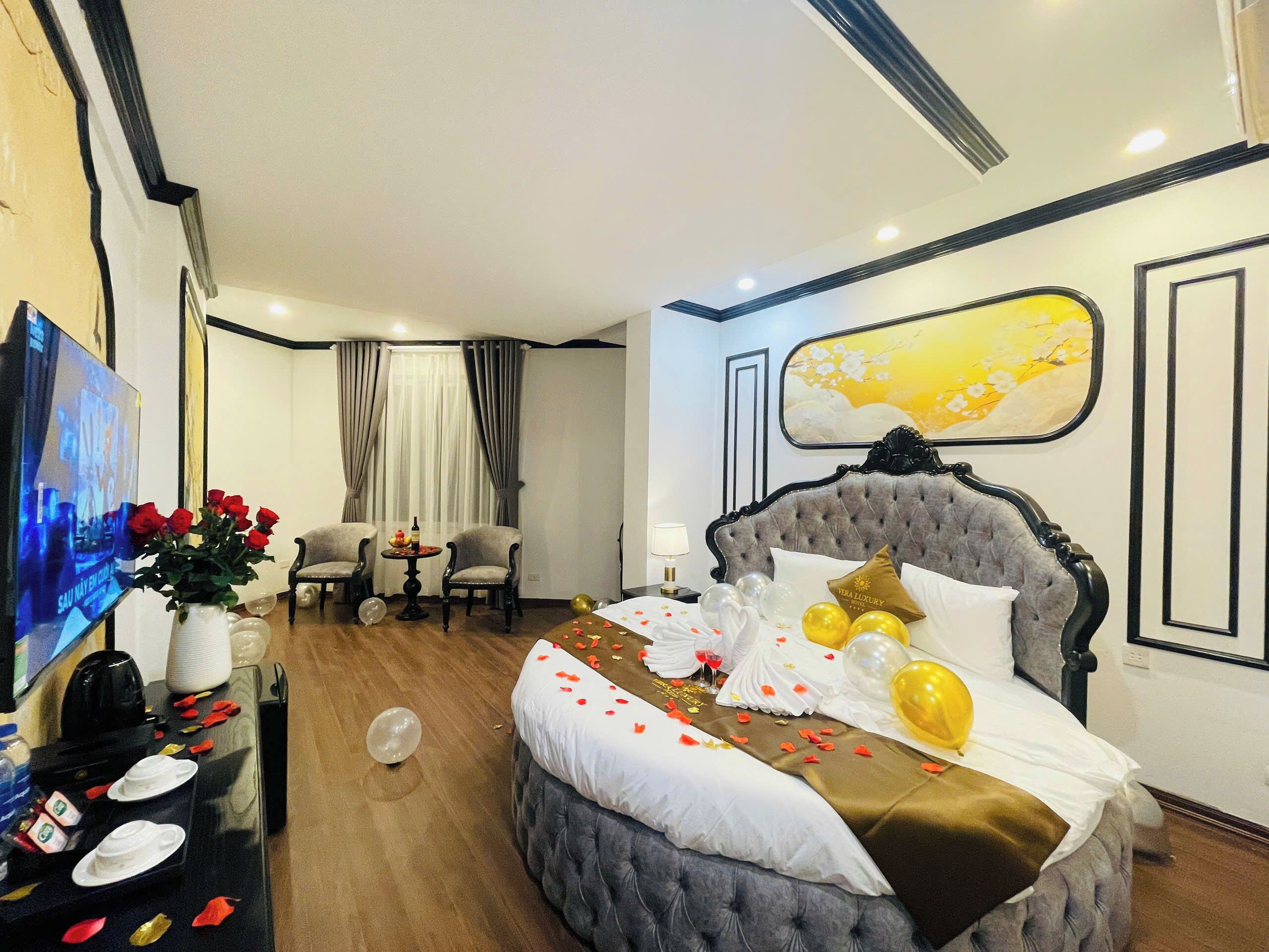 Una habitación de hotel con una cama grande, un televisor, un escritorio con un florero y una mesa de comedor con una botella y vasos.