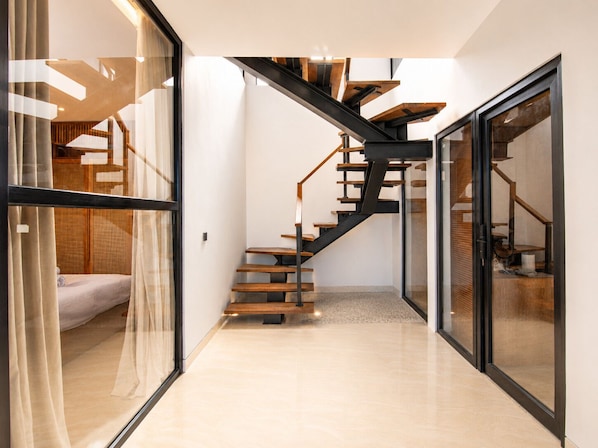 A modern interior with a glass door leading to a bedroom, a staircase with wooden steps and metal railings, and a tiled floor.