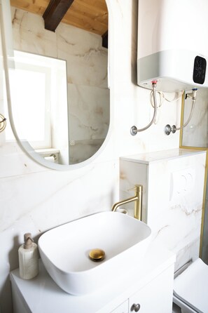 Un baño con lavamanos blanco, un espejo redondo, cabezal de ducha de mano y un calentador de pared.
