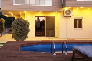 Une piscine avec une terrasse en bois, une porte vitrée et une fenêtre sur un bâtiment.