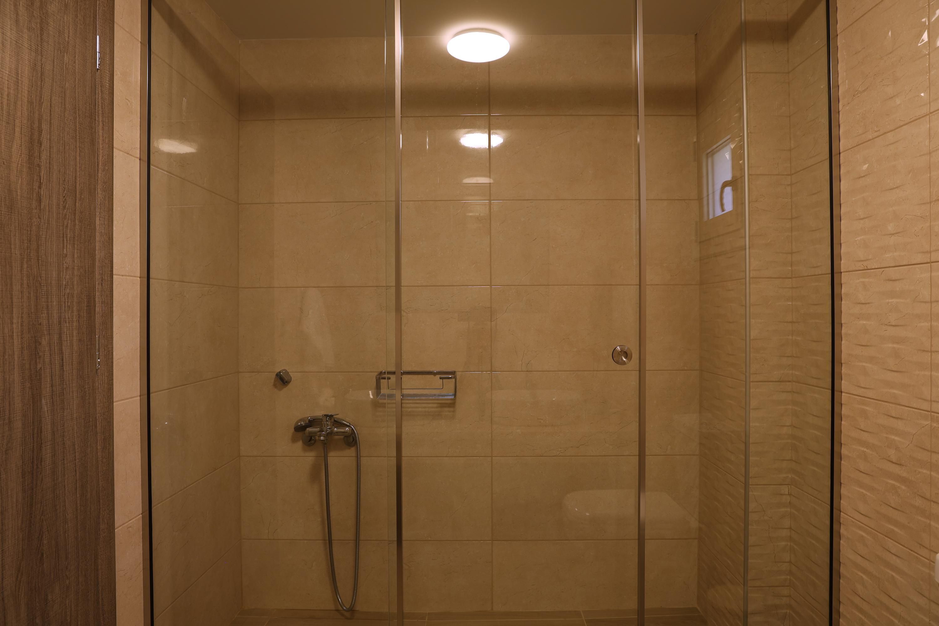 A glass-enclosed shower room with a tiled floor and walls, a handheld showerhead, and a control panel.