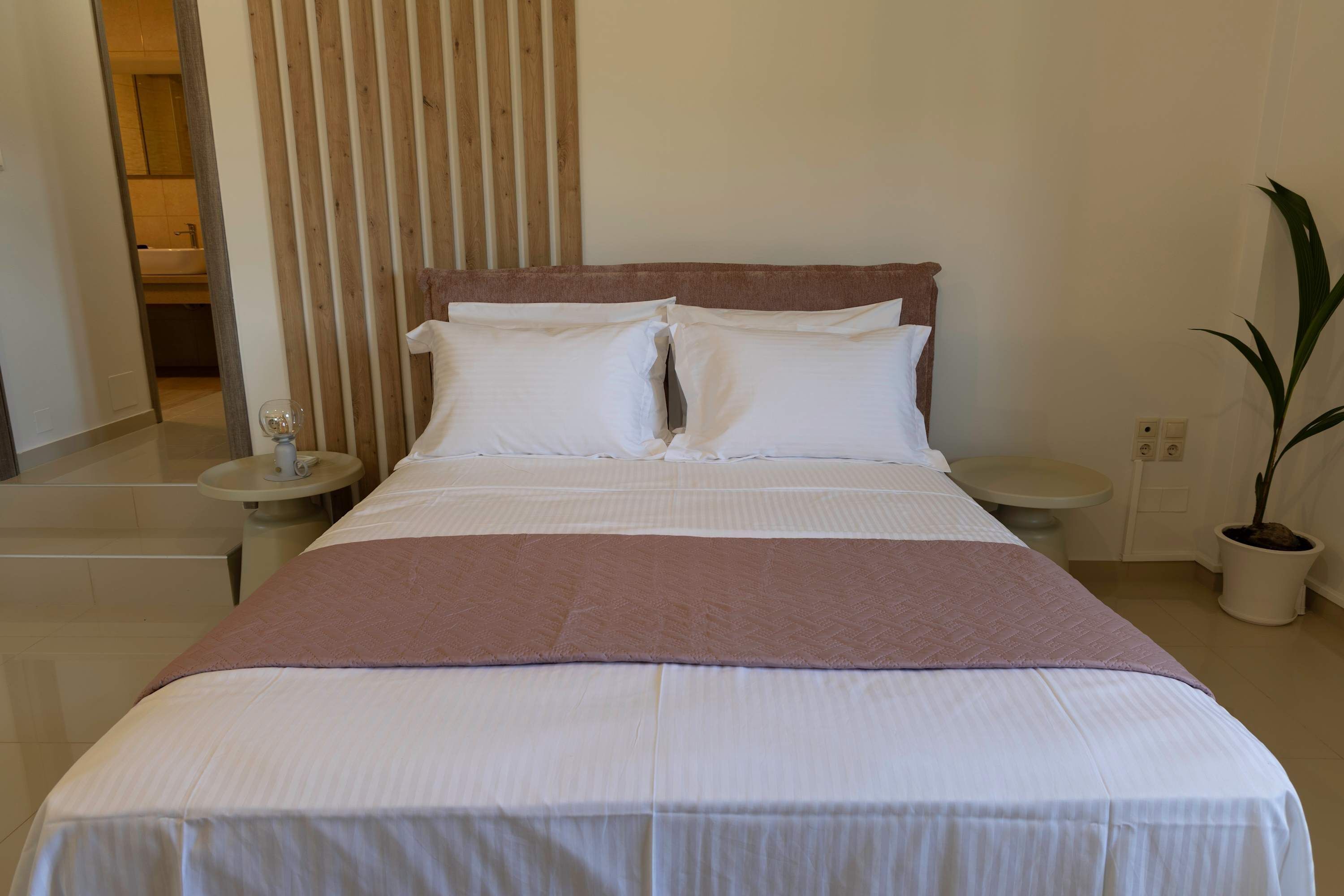 A neatly made bed with white and pink bedding, a wooden headboard, and a potted plant beside the bed.