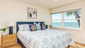 A bedroom with a bed, a ceiling fan, a window with a view of the ocean, and a nightstand with a lamp and a plant.