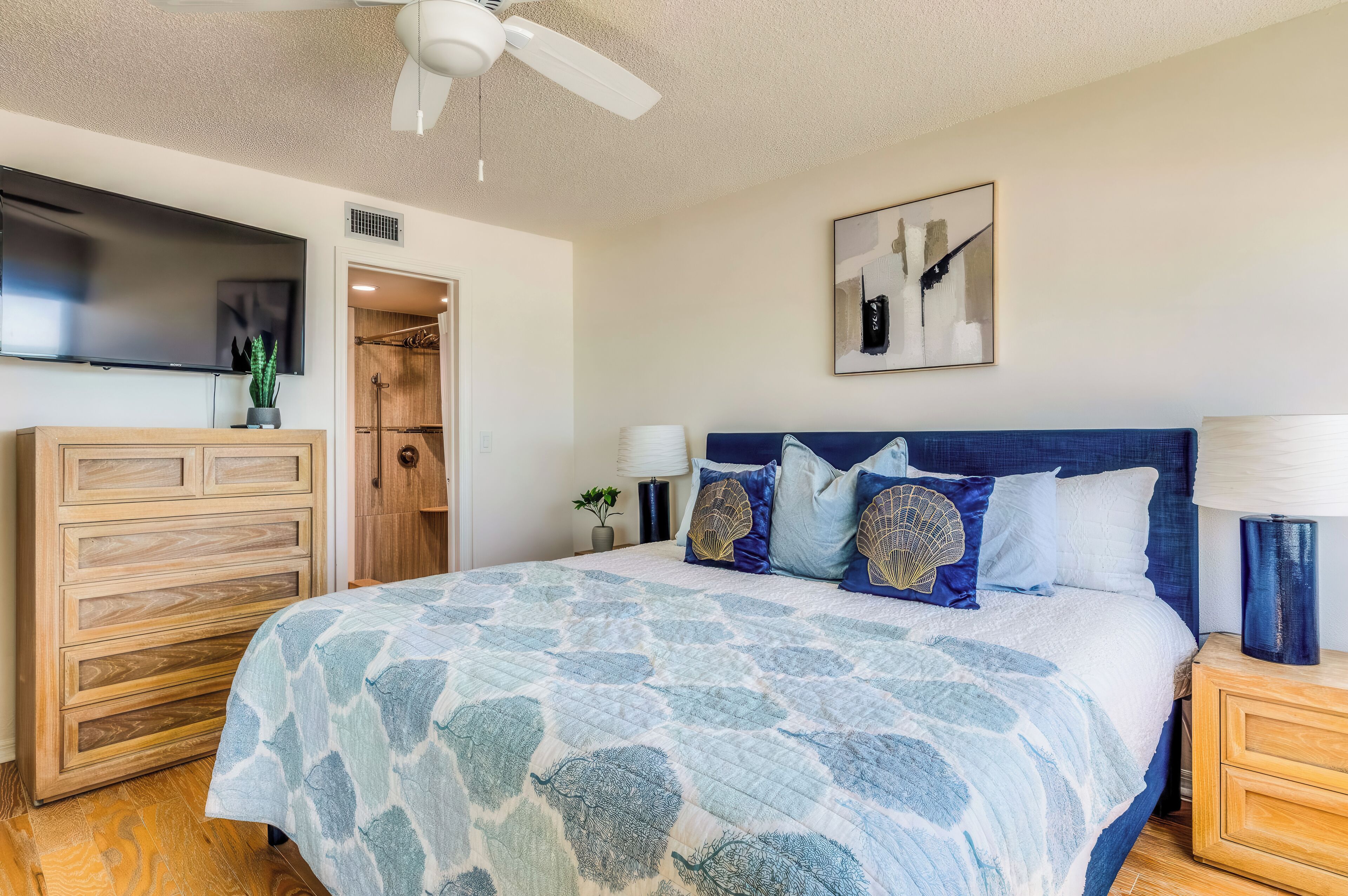 A bedroom with a bed, wooden dresser, ceiling fan, artwork, and a door leading to a bathroom.