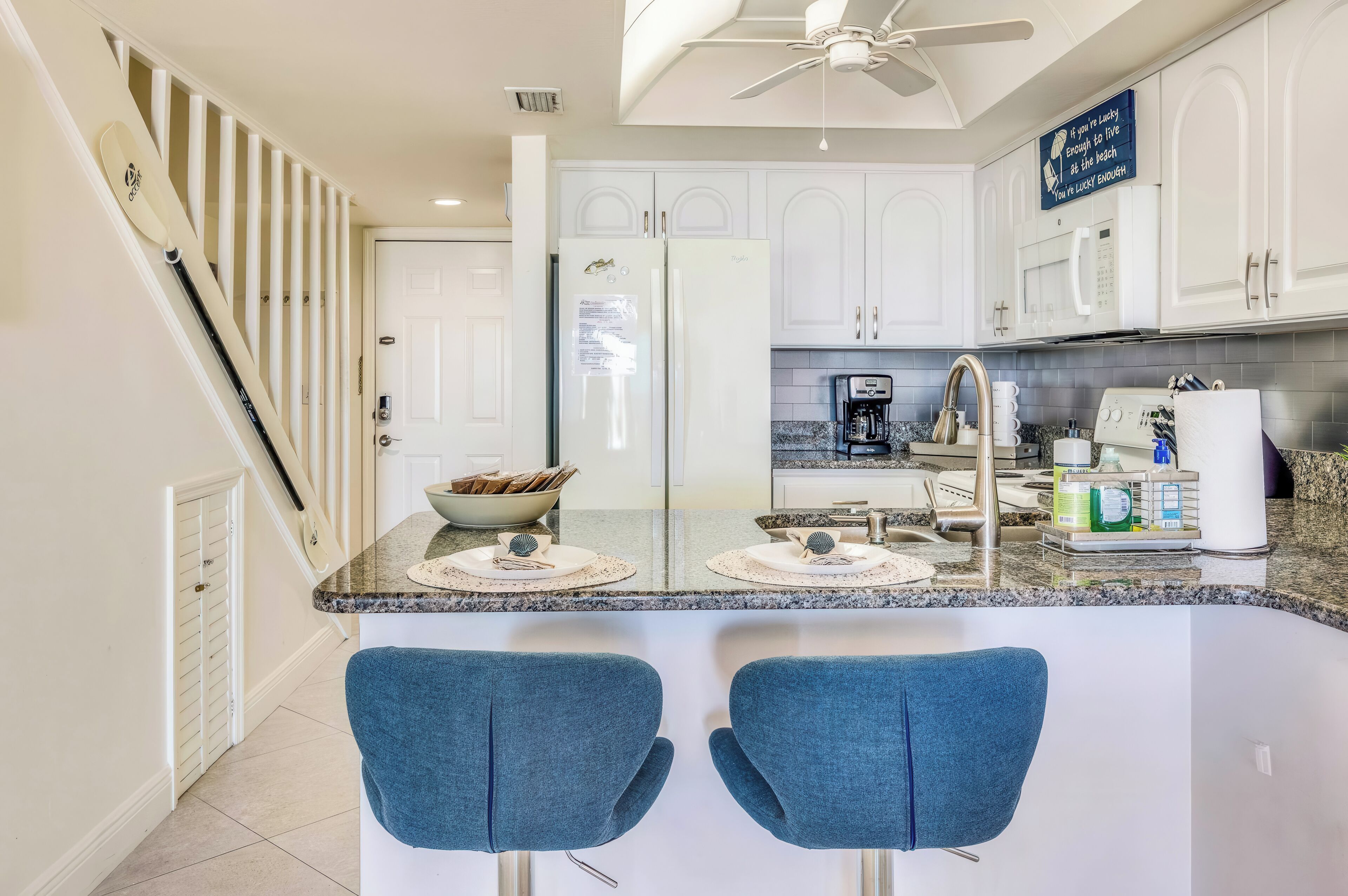 A modern kitchen with white cabinets, a central island with bar stools, a ceiling fan, and a sign on the wall.