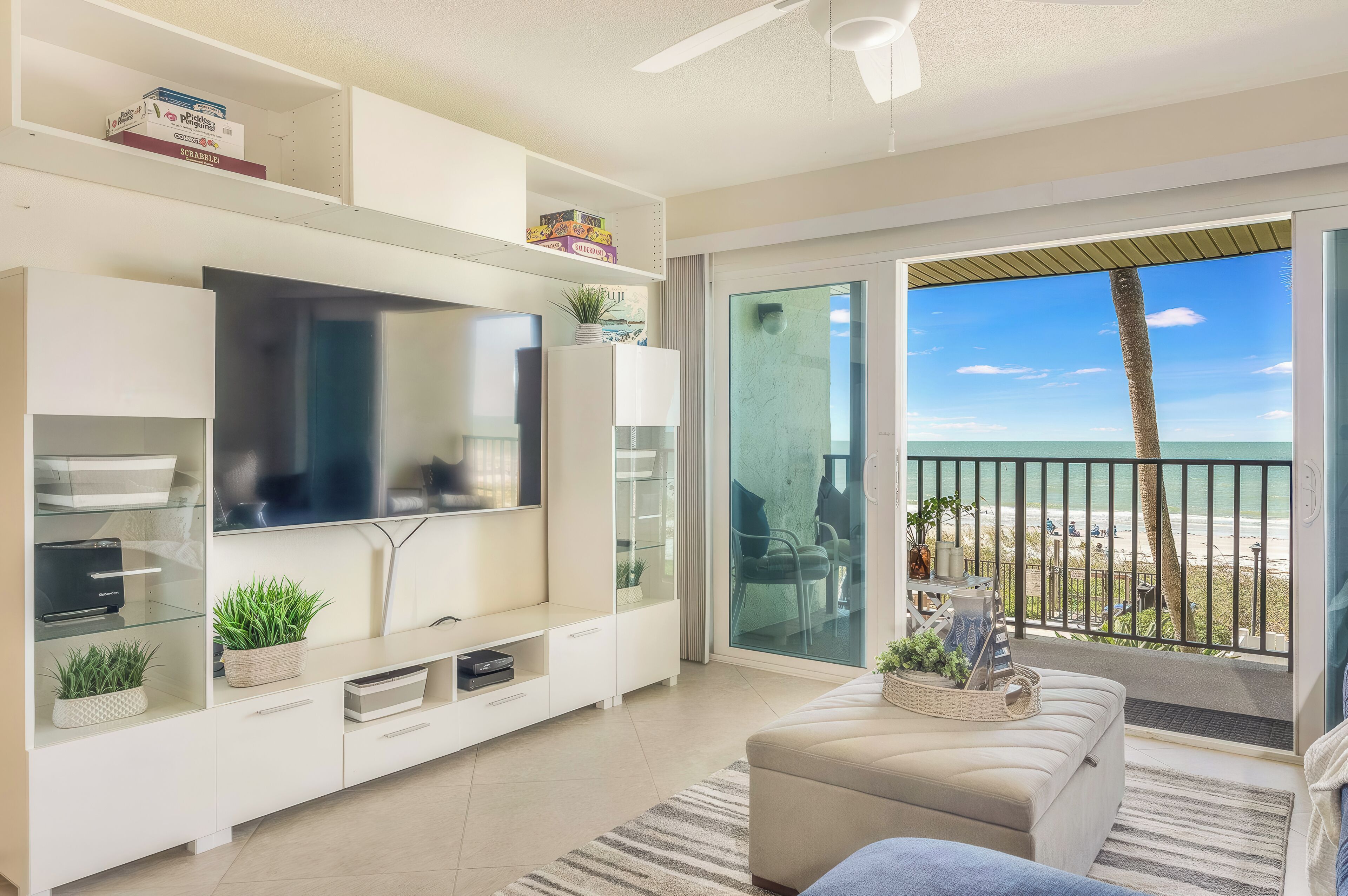 A modern living room with a flat-screen TV, a white entertainment unit, a sofa, and a balcony with a view of the beach.
