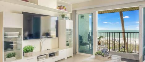 A modern living room with a flat-screen TV, a white entertainment unit, a sofa, and a balcony with a view of the beach.