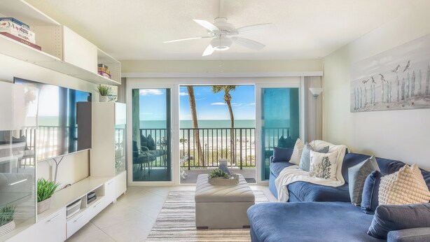 A living room with a TV, sofa, ottoman, and a view of the beach.