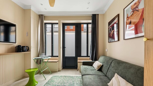 A modern living room with a green sofa, a small green table, a glass coffee table, and a large window with a view of buildings. - Charming Studio 2P Near Marais-bastille (Paris)