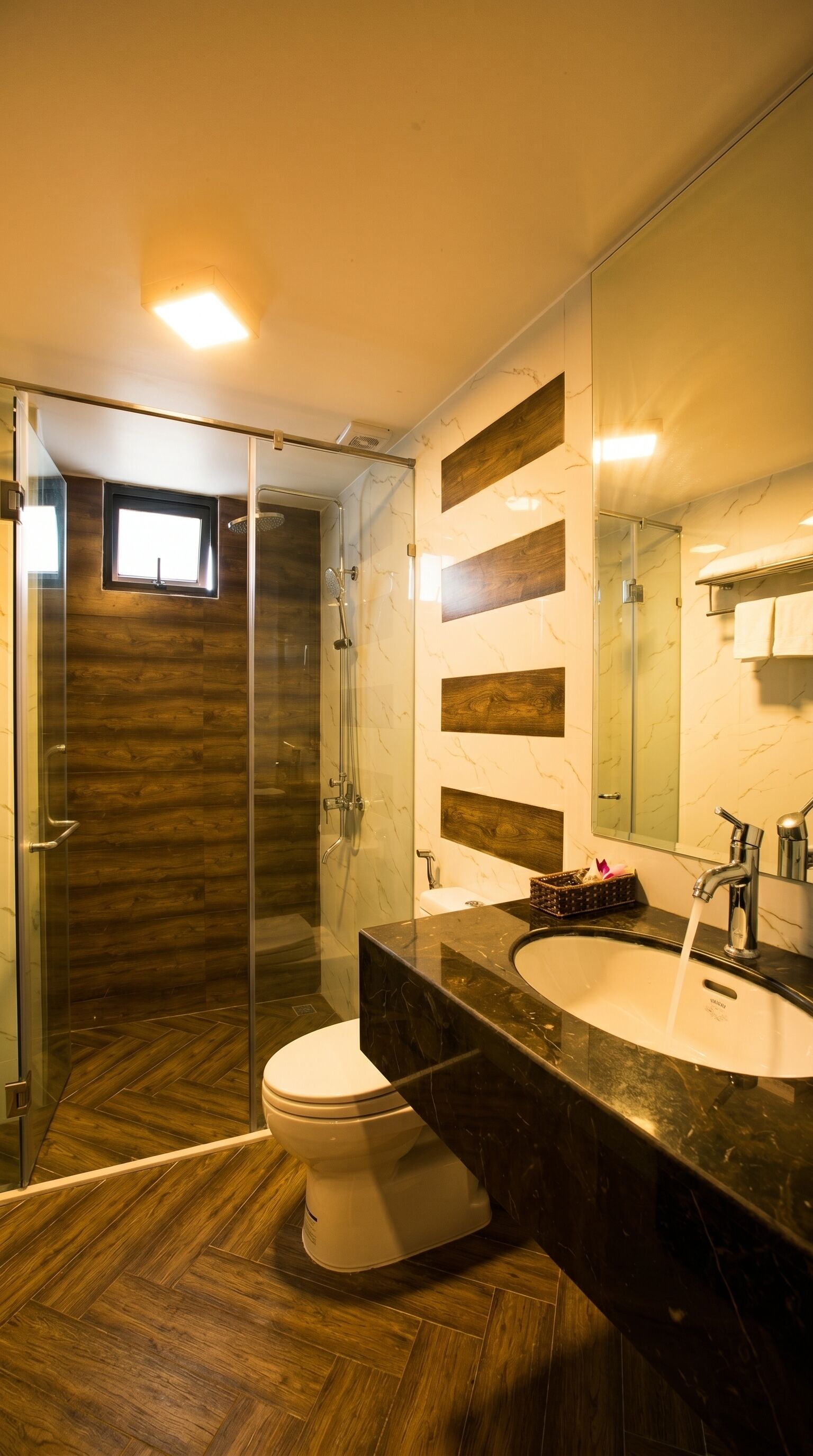 Une salle de bain moderne avec une douche à parois de verre, un plancher en bois, des toilettes blanches et un grand miroir au-dessus d’un lavabo en marbre foncé.