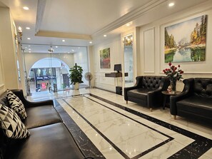 Um saguão de hotel com piso de mármore, sofás de couro preto, um balcão de recepção e uma grande pintura na parede.