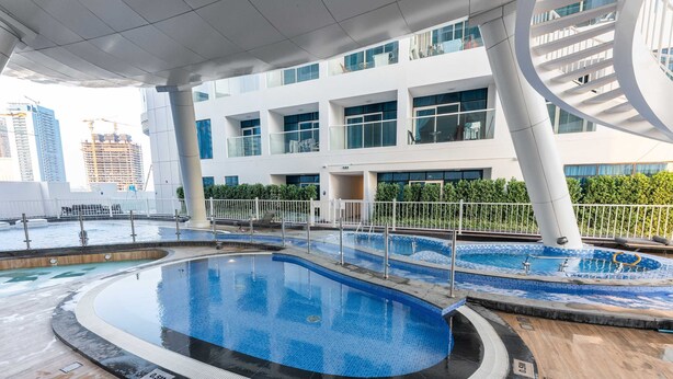 A pool area with multiple pools, a covered walkway, and a modern building with large windows in the background. - Free Housekeeping Visits During Your Stay - Stayshort - Stunning 2BR in Arjan That Sleeps 4 (Dubai)