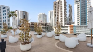 A rooftop terrace with potted plants, white seating, and a city skyline in the background. - Free Housekeeping Visits During Your Stay - Stayshort - 1BR in Jumeirah Village Circle Sleeps 4 With Balcony (Dubai)