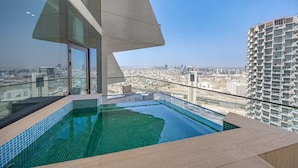 A rooftop infinity pool with a city view, a glass-enclosed balcony, and a modern building in the background. - Free Housekeeping Visits During Your Stay - Stayshort - 1BR in Jumeirah Village Circle Sleeps 4 With Balcony (Dubai)