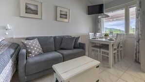Ein Zimmer mit grauem Sofa, einem kleinen weißen Tisch, einem an der Wand montierten Fernseher und einem Fenster mit Vorhängen.