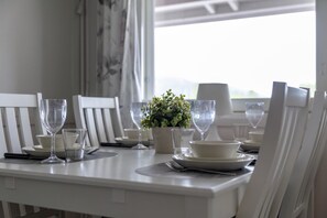 A dining table set with white chairs, plates, bowls, and glasses, with a potted plant and a window in the background. - Moonlight 402 | Private Sauna | Free Parking (Kittilä)