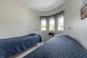Ein Zimmer mit zwei Betten, einem Vorhangfenster, einer Wandlampe und einem kleinen Regal.
