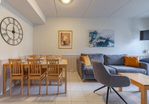 A dining area with a table and chairs, a grey sofa with orange cushions, and a painting on the wall. - Levistar 912 | 2BR | Sauna | Steps to Shops & Ski (Kittilä)