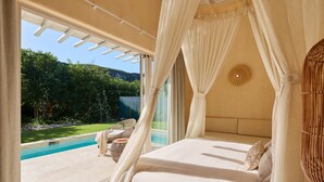 A bedroom with a bed, white curtains, a view of a swimming pool, and a wicker chair. - Luxury 7BR Villa With Private Pool in Cap Cana (Punta Cana)