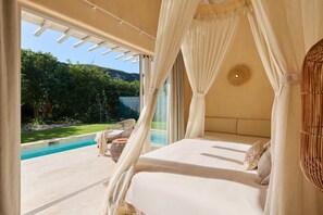 A bedroom with a bed, white curtains, a view of a swimming pool, and a wicker chair.