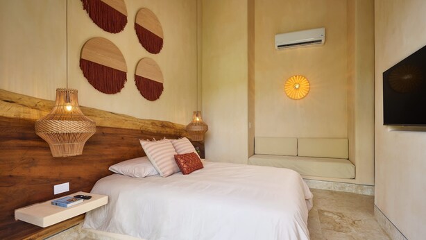 A bedroom with a wooden headboard, a bed with white bedding, a bedside table with a book and remote, wall-mounted decorative items, and a wall-mounted air conditioner. - Luxury 7BR Villa With Private Pool in Cap Cana (Punta Cana)