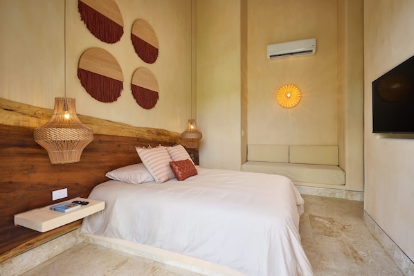 A bedroom with a wooden headboard, a bed with white bedding, a bedside table with a book and remote, wall-mounted decorative items, and a wall-mounted air conditioner.
