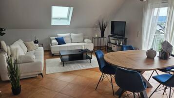 Ein modernes Wohnzimmer mit Sofa, Couchtisch, Esstisch, Stühlen, einem Fernseher und einem Fenster.
