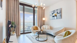 En moderne stue med sofa, lænestole, sofabord, fjernsyn og et stort vindue med gardiner.