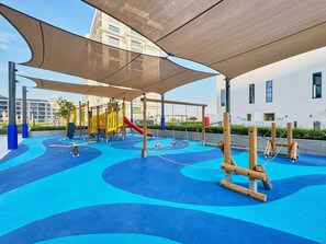 Ein Spielplatz mit Schaukeln, Rutschen und Klettergerüsten, umgeben von einer blauen Spielfläche und beschattet durch große Sonnensegel.