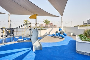 Ein Spielplatz mit Rutschen und Klettergerüsten, umgeben von blauer Bodenbelägung und beschattet durch große Sonnensegel.