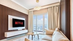 A living room with a flat-screen TV displaying 'NETFLIX,' a sofa, a small table, and a view of the city through large windows. - 2 Bedroom Apartment Close to Miracle Tower (Dubai)