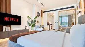 A modern hotel room with a large bed, a flat-screen TV, a plant, and a view of the city. - Luxury Studio in Trillionaire Residence (Dubai)