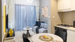 Ett kompakt bostadsutrymme med en matplats, en soffa, ett litet bord, ett kök med en gasspis och ett stort fönster med tunna, genomskinliga gardiner.
