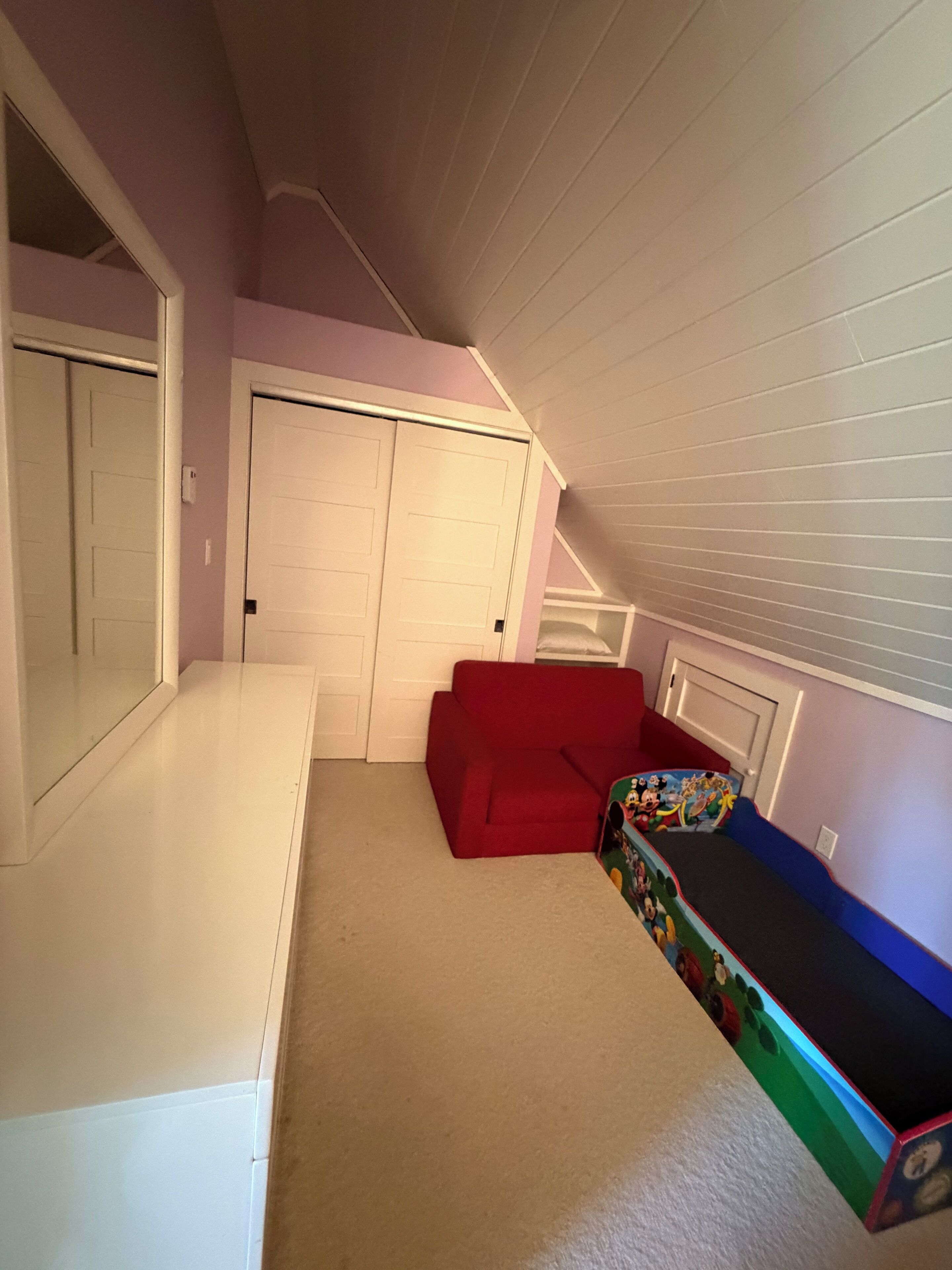 Une pièce avec un plafond en pente, un canapé rouge, une commode blanche et une porte.