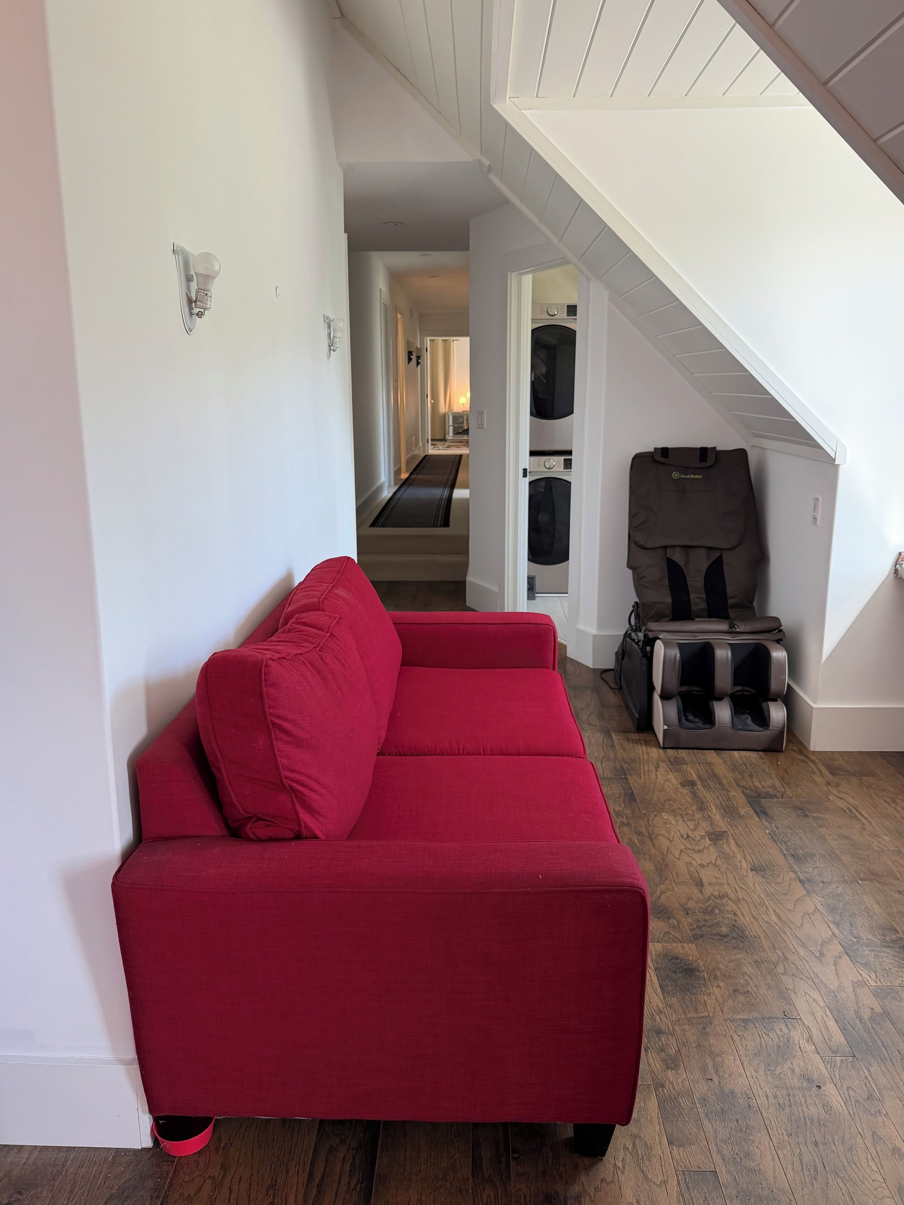 Ein rotes Sofa in einem Raum mit Holzboden, einer Treppe und einem an der Wand befestigten Massagesessel.