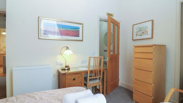 A bedroom with a bed, a desk with a lamp, a chair, a wardrobe, and a door leading to another room. - Pincerna Apartment Suite 1a (Aberdeen)