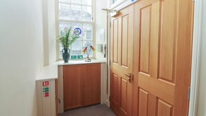 A hallway with a wooden door, a window with a view of buildings, a vase with flowers, and a fire exit sign. - Pincerna Apartment Suite 1a (Aberdeen)