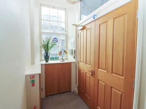 Un couloir avec une porte en bois, une fenêtre donnant sur des immeubles, un vase rempli de fleurs et un panneau indiquant la sortie de secours.