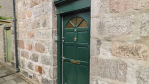 A stone building with a green door, a visible window, and a small outdoor area with a green door and a wall-mounted device. - Pincerna Apartment Suite 1a (Aberdeen)