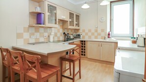 A kitchen with wooden cabinets, a central island with stools, a window with a view, and a clock on the wall. - Pincerna Apartment Suite 1a (Aberdeen)
